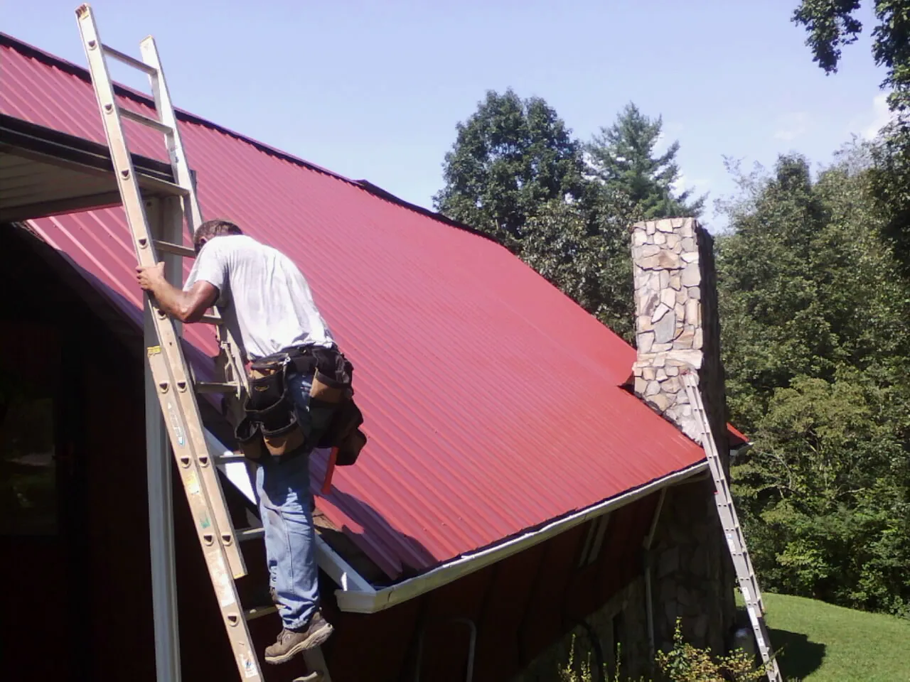 Roofer on a ladder beside a red metal roof and stone chimney — Soffit & Fascia Repair by Roofing Brothers in Hooppole